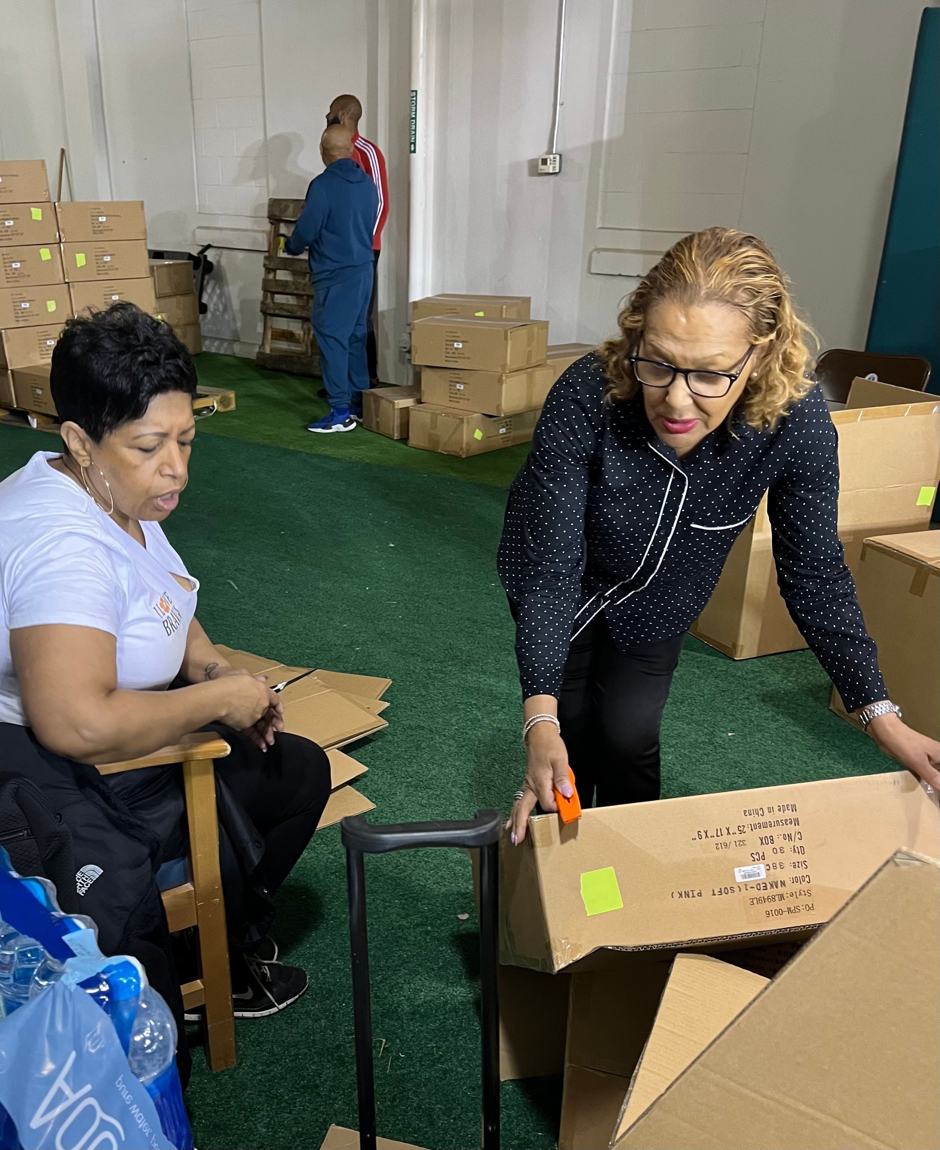 Two women are standing next to each other in a room filled with cardboard boxes.