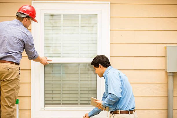 Two professionals in work clothes inspect a window on the exterior of a tan building; one wears a red safety helmet.