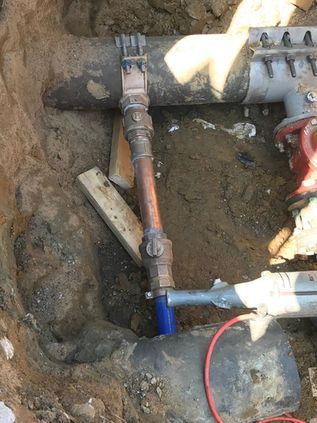 Utility pipes and valves exposed in a dirt trench, with copper and blue piping connected underground.
