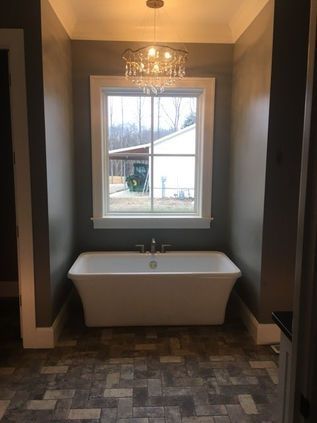 Modern bathroom with a freestanding tub under a window, dark walls, patterned tile floor, and chandelier lighting