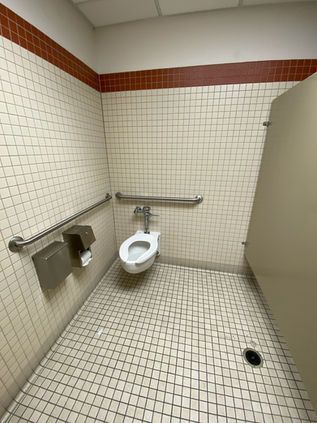 Accessible restroom stall with wall-mounted toilet, grab bars, tiled walls, and floor drain.