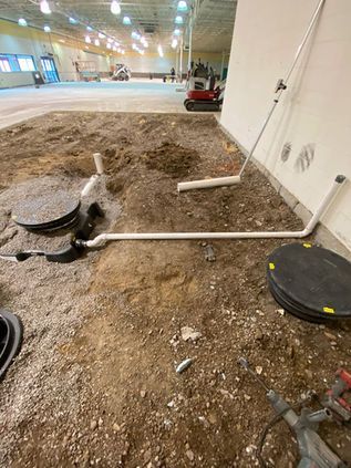 Indoor construction area with dirt floor and scattered pipes, cables, and round equipment under bright lights
