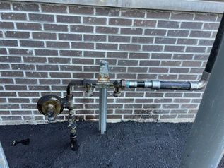 Utility pipes and valves mounted against a brick wall on a gravel surface.