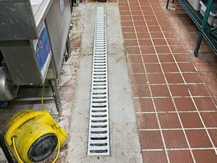 Concrete floor drain with a long metal grate beside terracotta tiles and kitchen equipment.