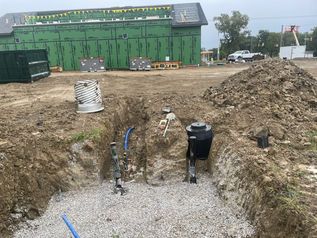Construction trench with exposed pipes and gravel beside a green building and dirt piles