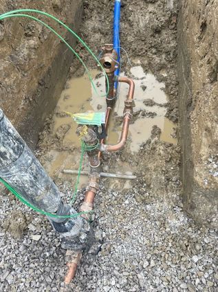 Open trench with exposed copper pipes, blue conduit, and utility equipment in gravel soil.