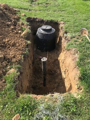 Excavation trench in grass with a black cylindrical tank and a central post or pipe exposed underground