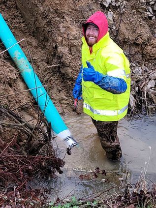 Worker in neon rain gear stands in muddy water beside a blue pipe at a construction site.