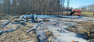Construction site with foundation work, workers, and an excavator on a cleared lot near trees