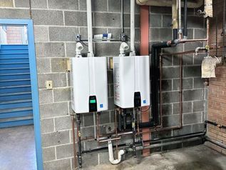 Two white wall-mounted water heaters in a utility room with exposed pipes and cinderblock walls
