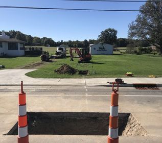 Road construction site with an open trench, orange cones, and an excavator on a grassy roadside.