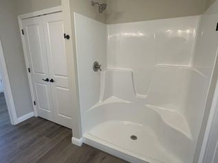 Bathroom shower with white tiled surround and gray wood-look flooring beside a white door.