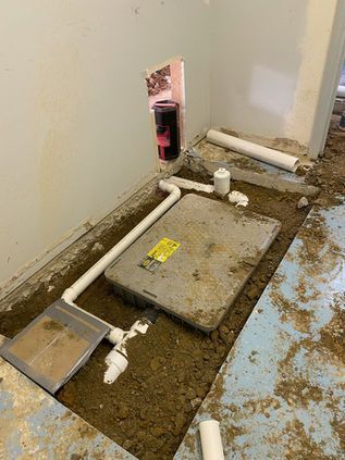 Dirty utility room floor with an open plumbing access panel and pipes exposed.