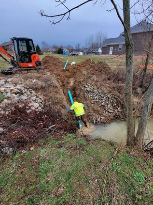 Worker in a neon jacket installs blue pipe in a muddy trench beside an orange excavator.