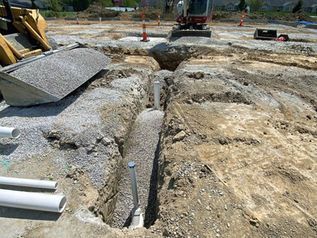 Construction site trench with exposed pipes, gravel, and heavy equipment in the background