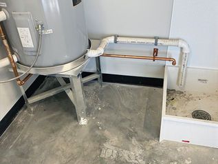 Utility room with large gray tank, exposed pipes, and a white floor drain basin on a concrete floor