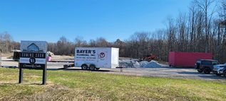 Rural roadside with a Bayer’s trailer, red truck, and piles of materials near a wooded lot