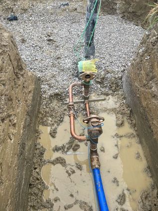 Trench with exposed plumbing pipes, valves, and a blue hose in muddy standing water