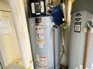 Utility room with two water heaters, pipes, and a blue tool hanging on the wall.