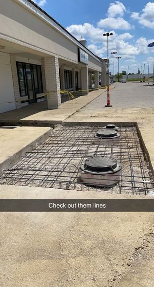 Building walkway with exposed rebar and round utility covers beside a concrete sidewalk under a blue sky