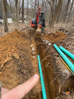 Excavator digging a trench in a wooded area beside blue pipes; a finger points at the trench.
