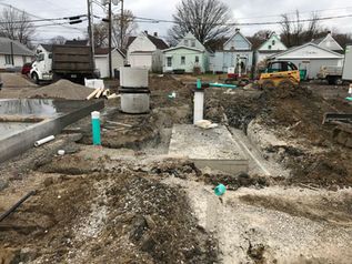 Muddy construction site with concrete foundations, blue pipes, and houses in the background