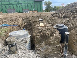 Excavation site with buried tanks and pipes in muddy trench beside a green building