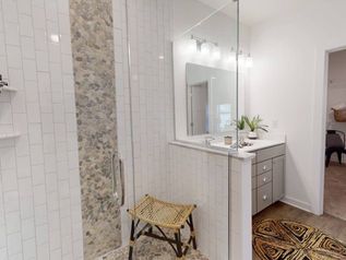 Bright bathroom with glass shower, white vanity, and wicker stool on patterned floor rug.