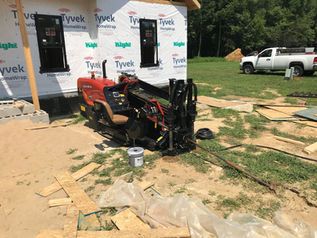 Red skid steer beside a house under construction, with plywood, tools, and a pickup truck nearby.