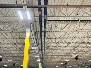 Warehouse ceiling with metal trusses, bright lights, and a yellow support beam underneath.
