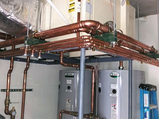 Copper pipes and two gray water heaters in a utility room with exposed ceiling piping