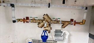 Plumbing and water meter assembly with brass valves and blue filter in a utility alcove