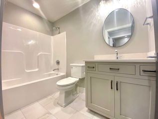 Bright bathroom with white tub, toilet, gray vanity, and round mirror