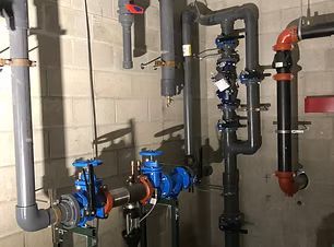 Industrial pipes and valves in a utility room, with blue pumps and red fittings against cinderblock walls