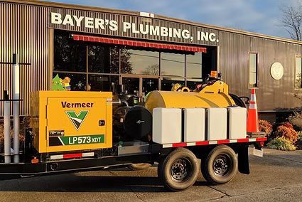 Yellow Vermeer trailer parked outside Bayer's Plumbing, Inc. building with work equipment loaded on top