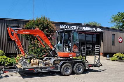 Orange excavator on a trailer outside Bayer’s Plumbing, Inc. storefront in bright daylight