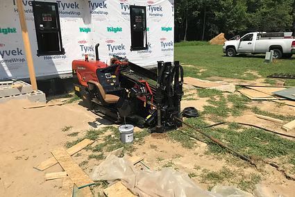 Red and black skid steer loader parked on a concrete pad beside a Tyvek-wrapped building, with tools and lumber nearby