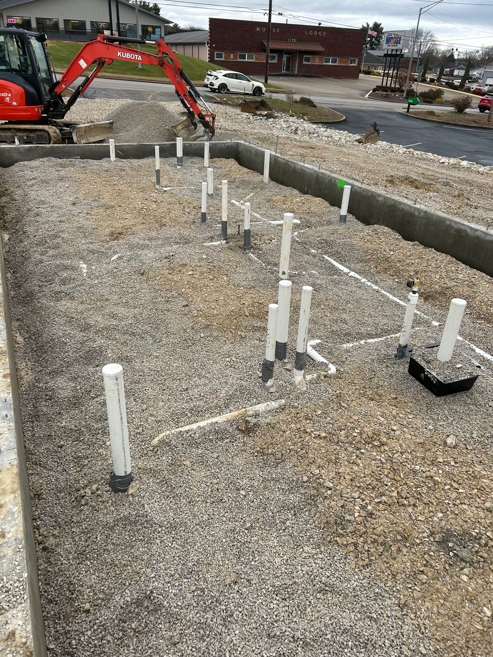 Construction site with gravel foundation, white PVC pipes, and an orange excavator in the background