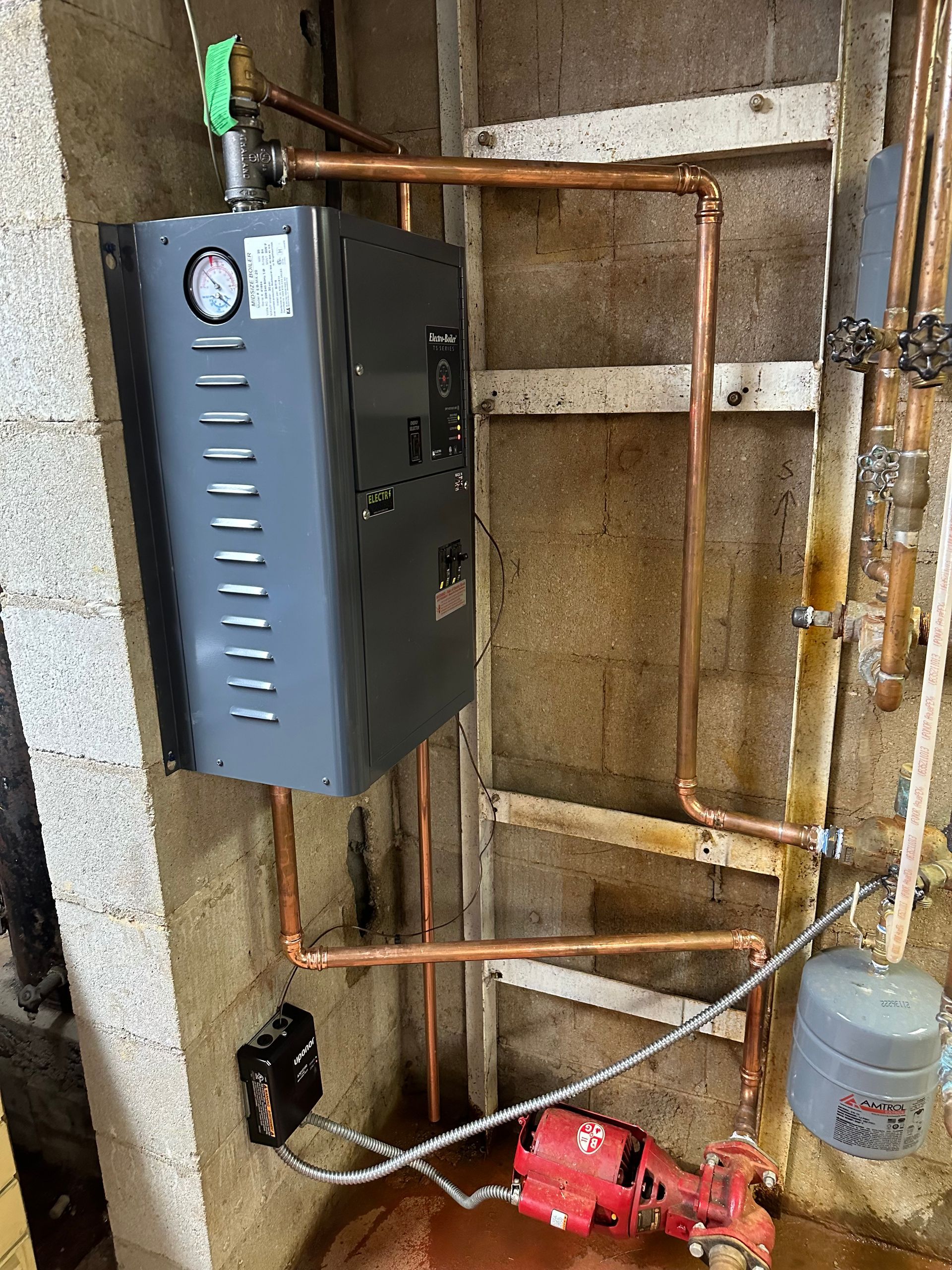 Basement boiler setup with wall-mounted control box, copper pipes, and a red pump on the floor
