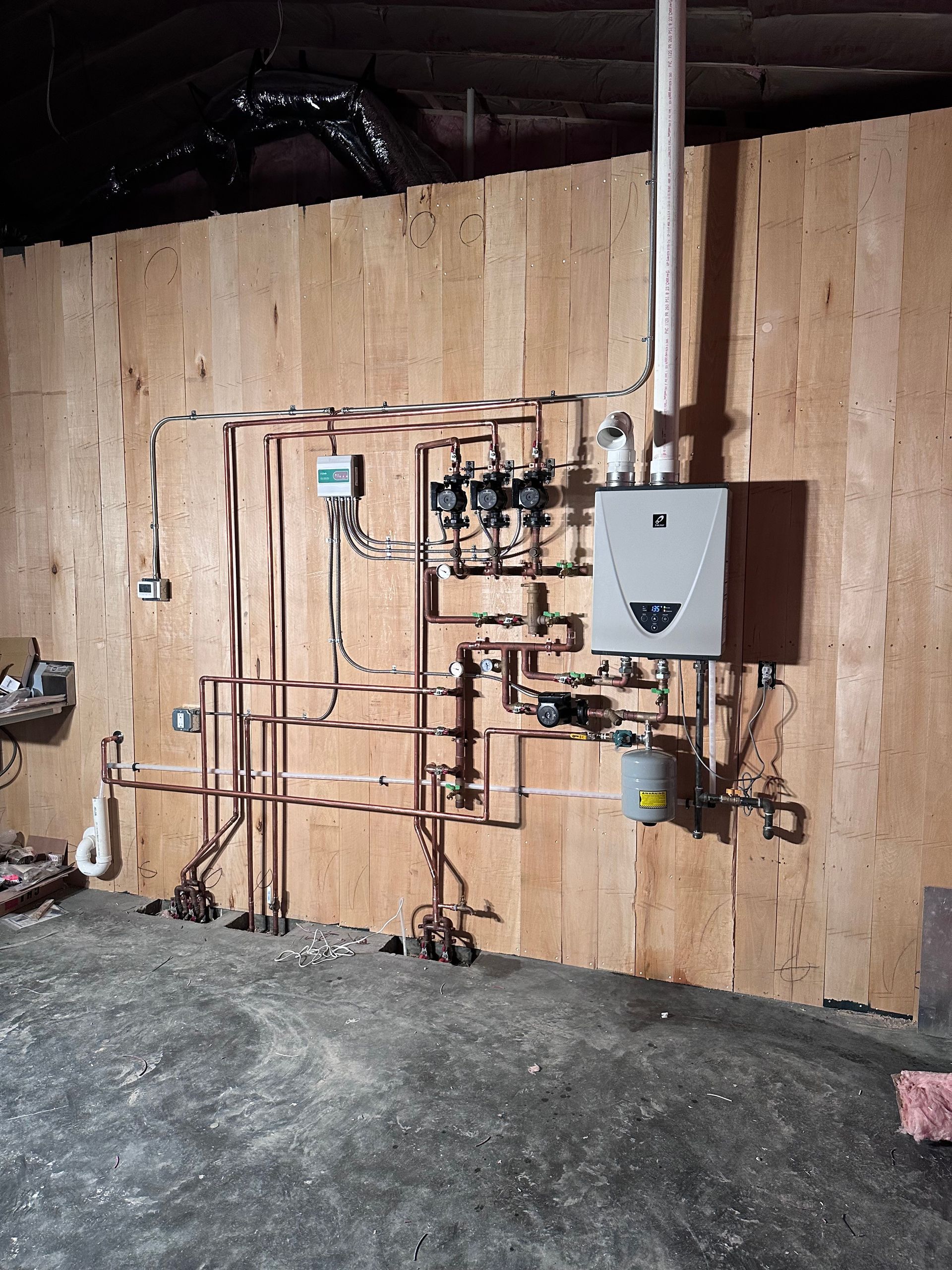 Wall-mounted plumbing and heating pipes on a plywood wall in a basement or utility room.