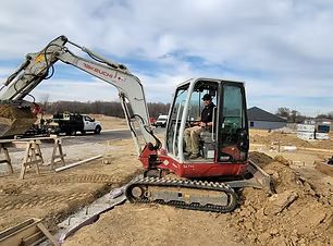 Small excavator digging a trench at a construction site on a dirt road, with a worker in the cab.