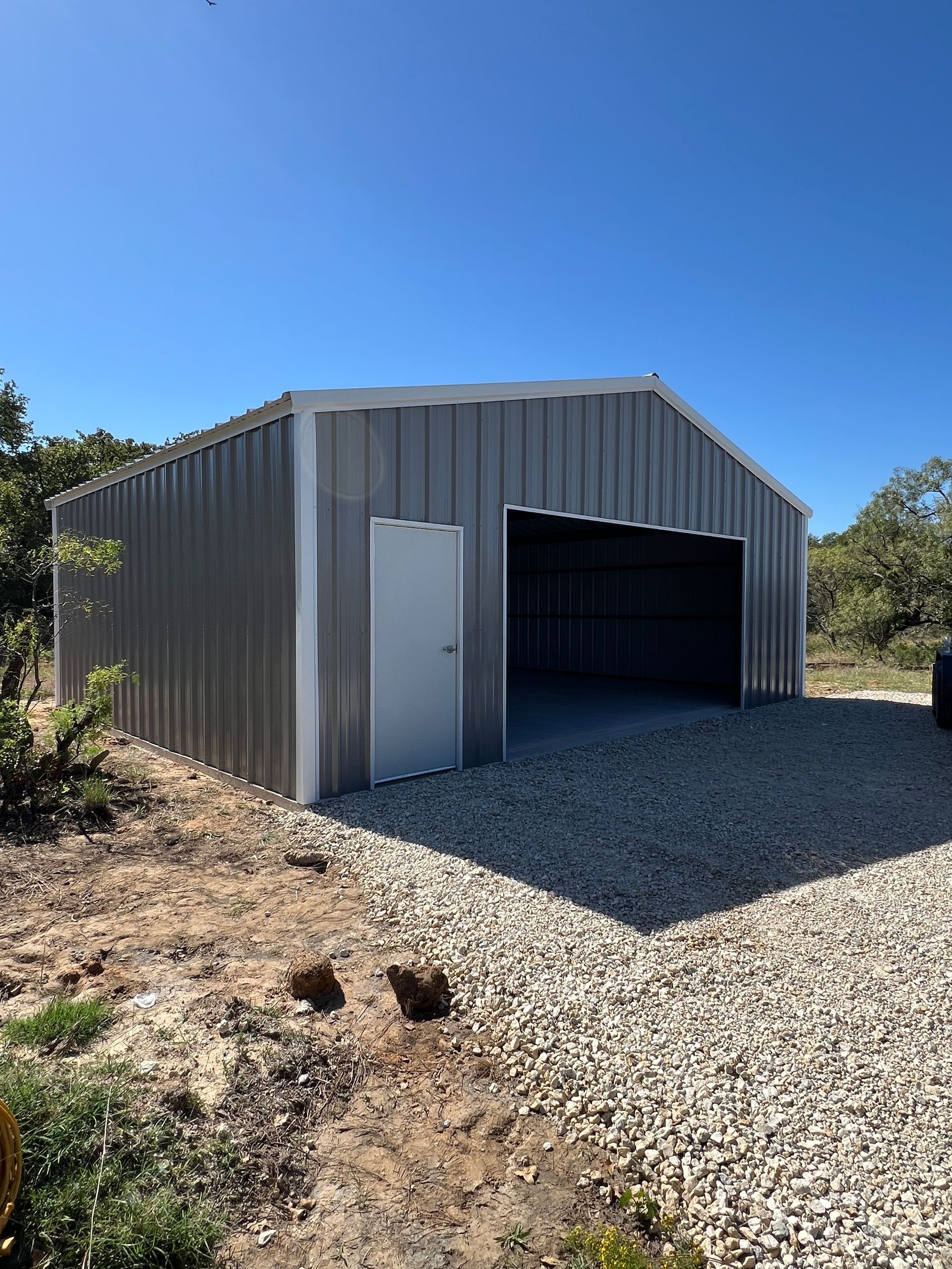 Gray metal shed with open bay door and white side door on a gravel lot under a clear blue sky