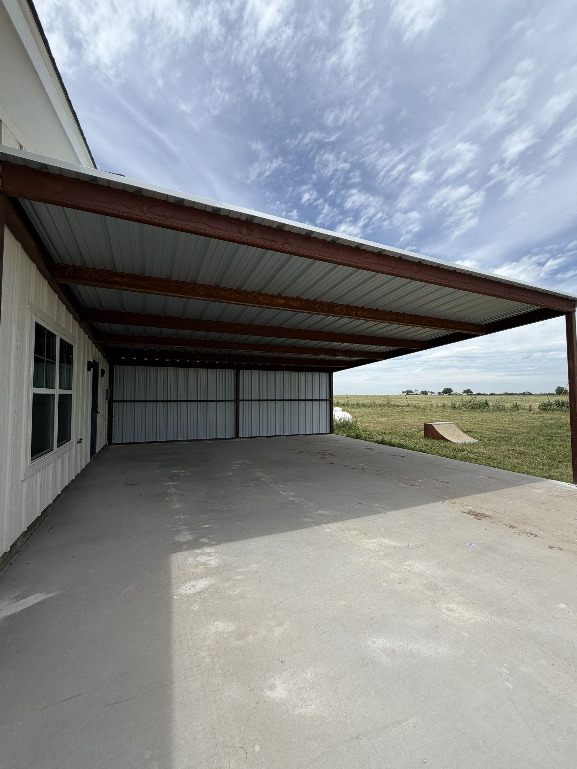 Covered concrete patio beside a white building, with a metal roof and open grassy field beyond.