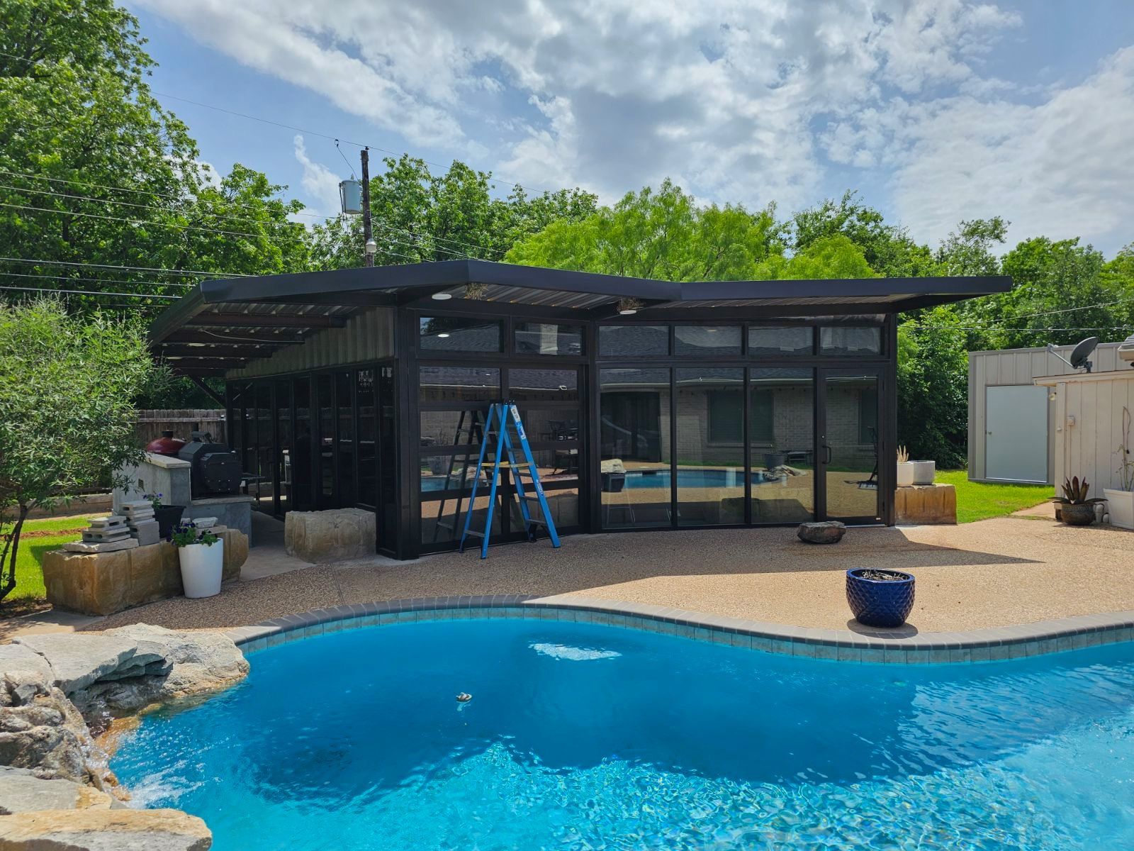 Backyard pool beside a black glass cabana with a blue ladder and potted plants