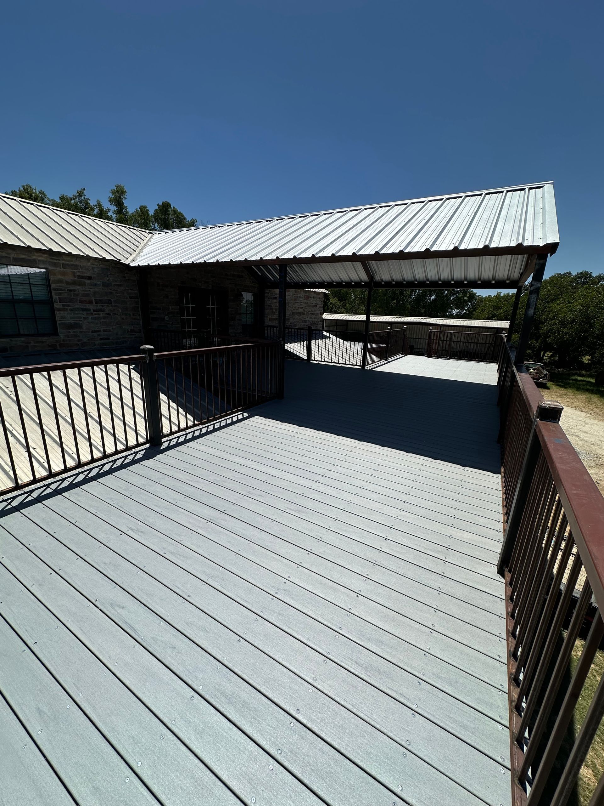 Rooftop deck with light gray metal siding and railings, connected to a rustic house under a blue sky.