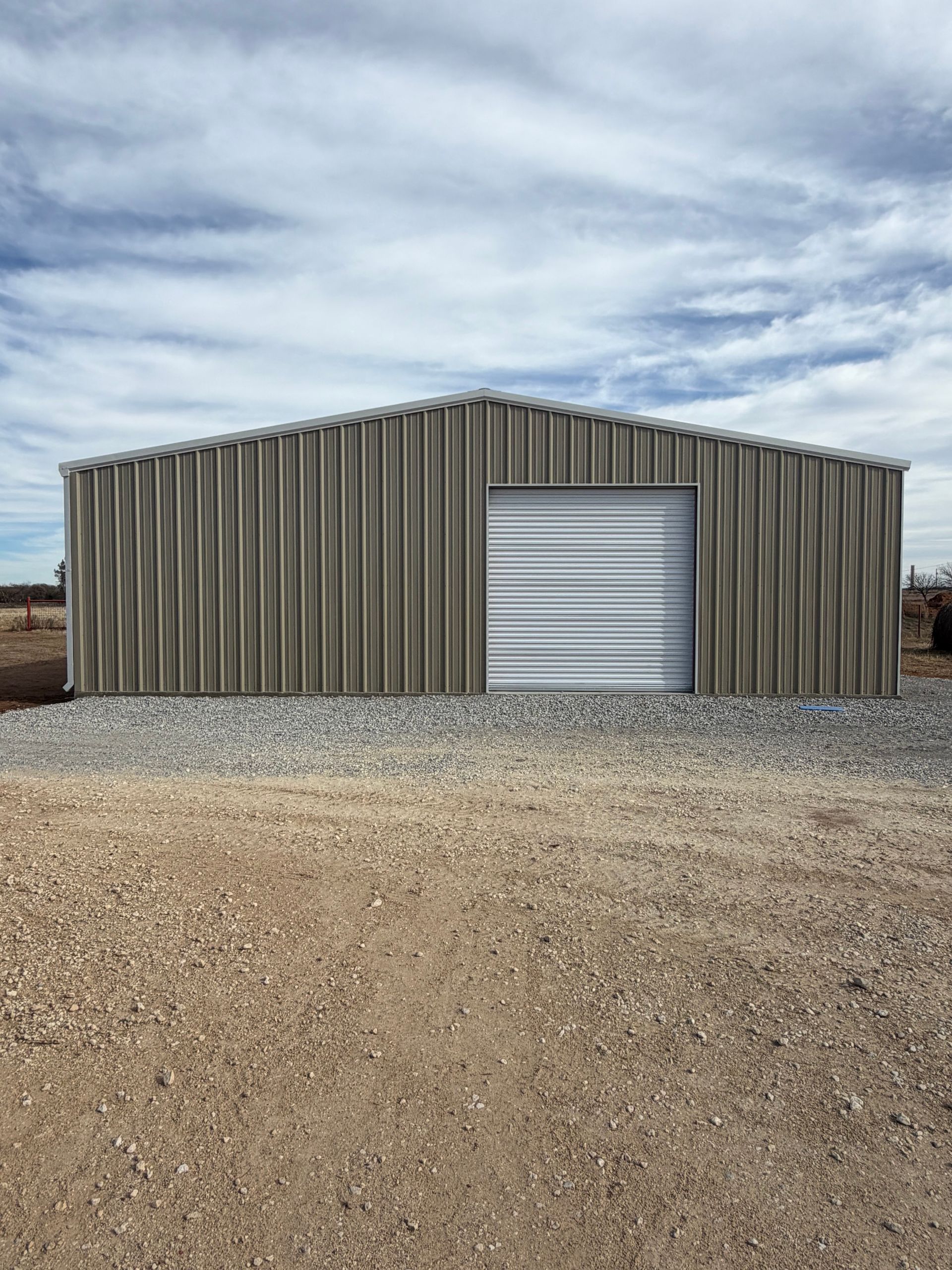 Gray metal storage shed with a roll-up door on a gravel lot under a cloudy sky