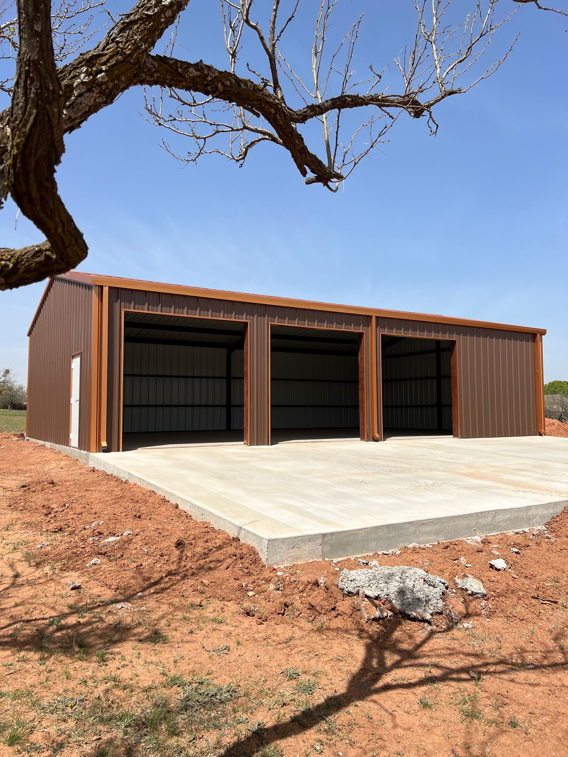 Rust-colored metal garage with three black doors on a concrete pad in a dry, open landscape.