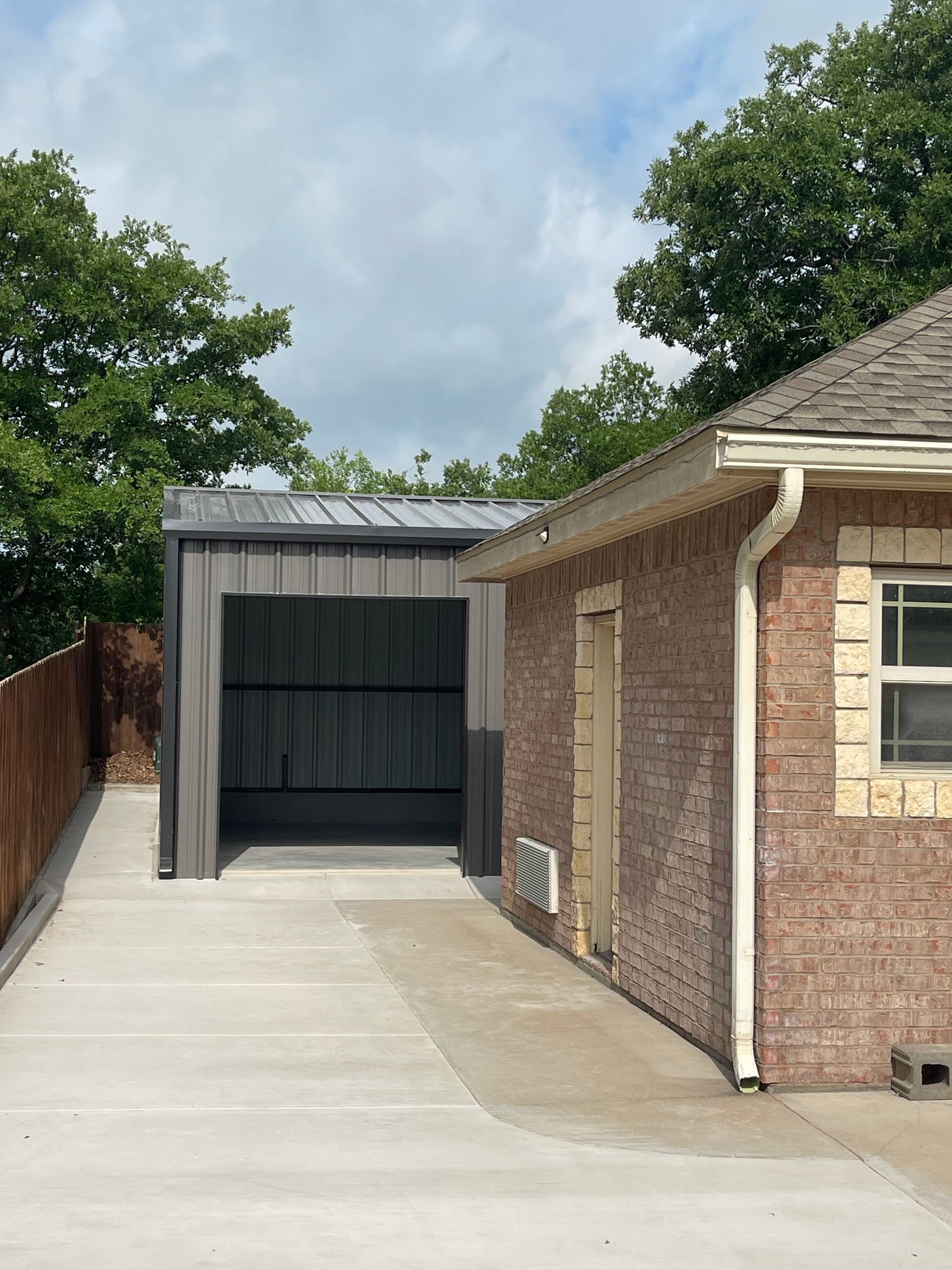Concrete driveway beside a brick house leads to a covered carport and wooden fence under a partly cloudy sky