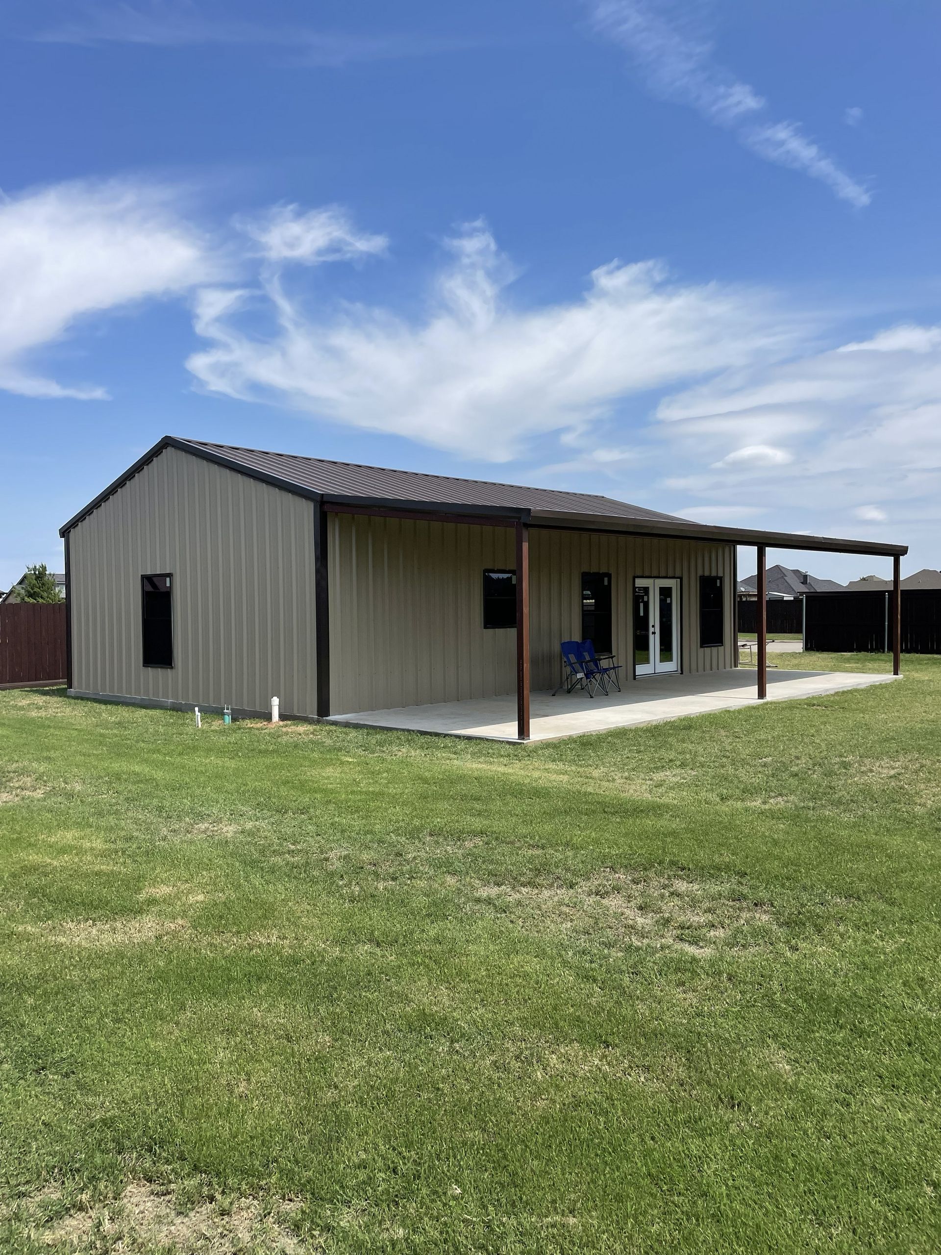 Small tan metal house with a porch and chairs, set in a grassy yard under a blue sky.