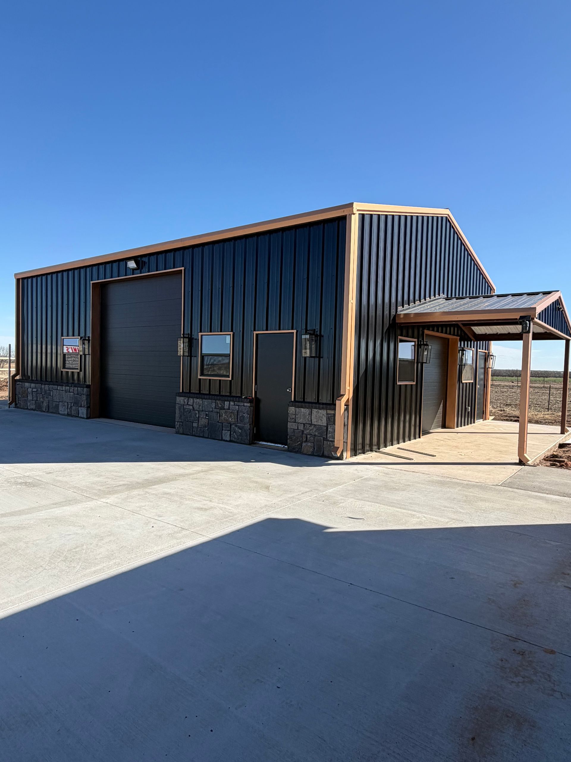 Rustic black metal building with a covered entrance and stone base in a sunny open lot.
